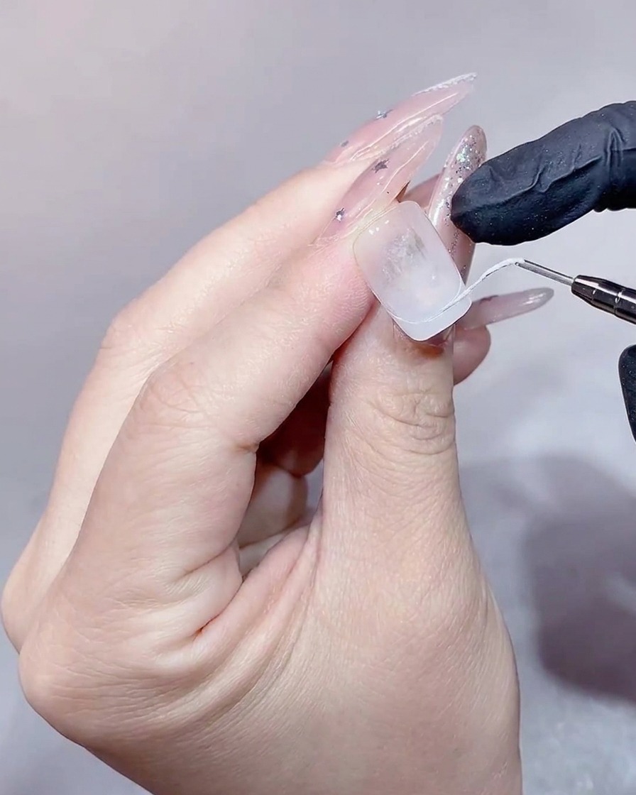 Primer plano de un manicurista utilizando un pincel plano para rellenar una punta francesa blanca en una extensión de uña transparente.