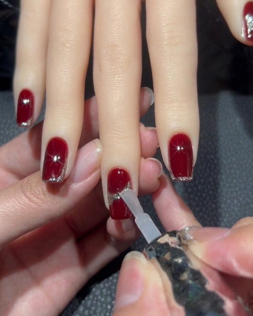 Un pincel aplicando top coat transparente sobre una uña roja con un diseño de glitter, rodeando con cuidado una piedra brillante central.