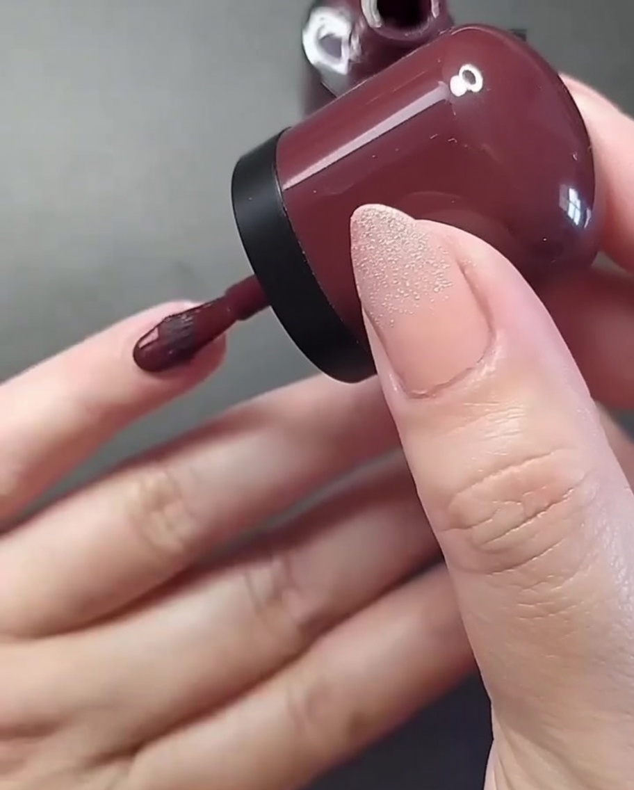 Primer plano de una mano con un pincel aplicando esmalte burdeos intenso en la uña del dedo meñique.