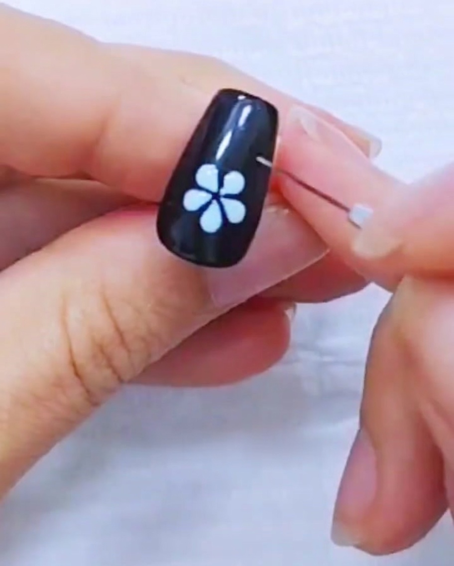 Primer plano de un pincel liner refinando los bordes de un diseño de flor blanca de cinco pétalos sobre una uña negra.