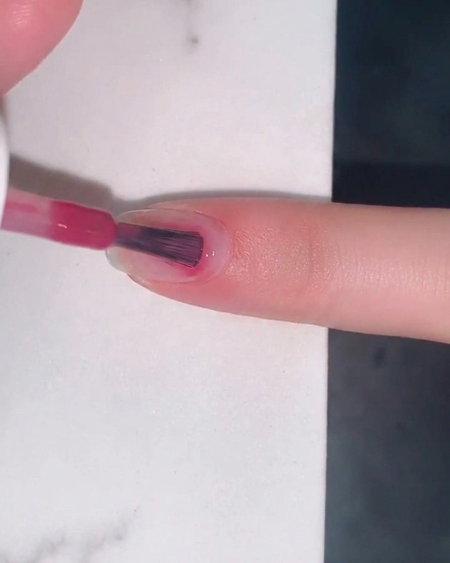 Un pincel aplicando una capa translúcida de esmalte de gel jelly color rosa baya en una uña natural.