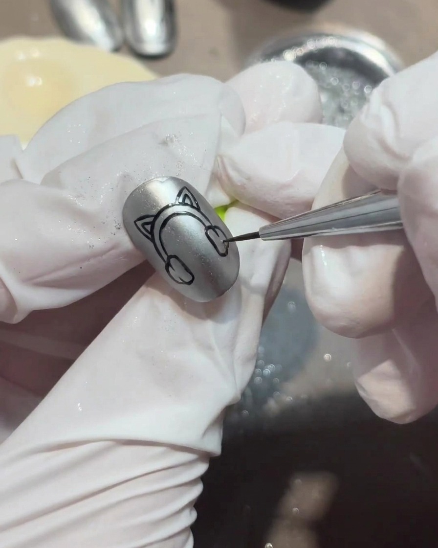 Primer plano de una mano enguantada usando un pincel fino para pintar formas de orejas de gatito con top coat brillante sobre una uña cromada.