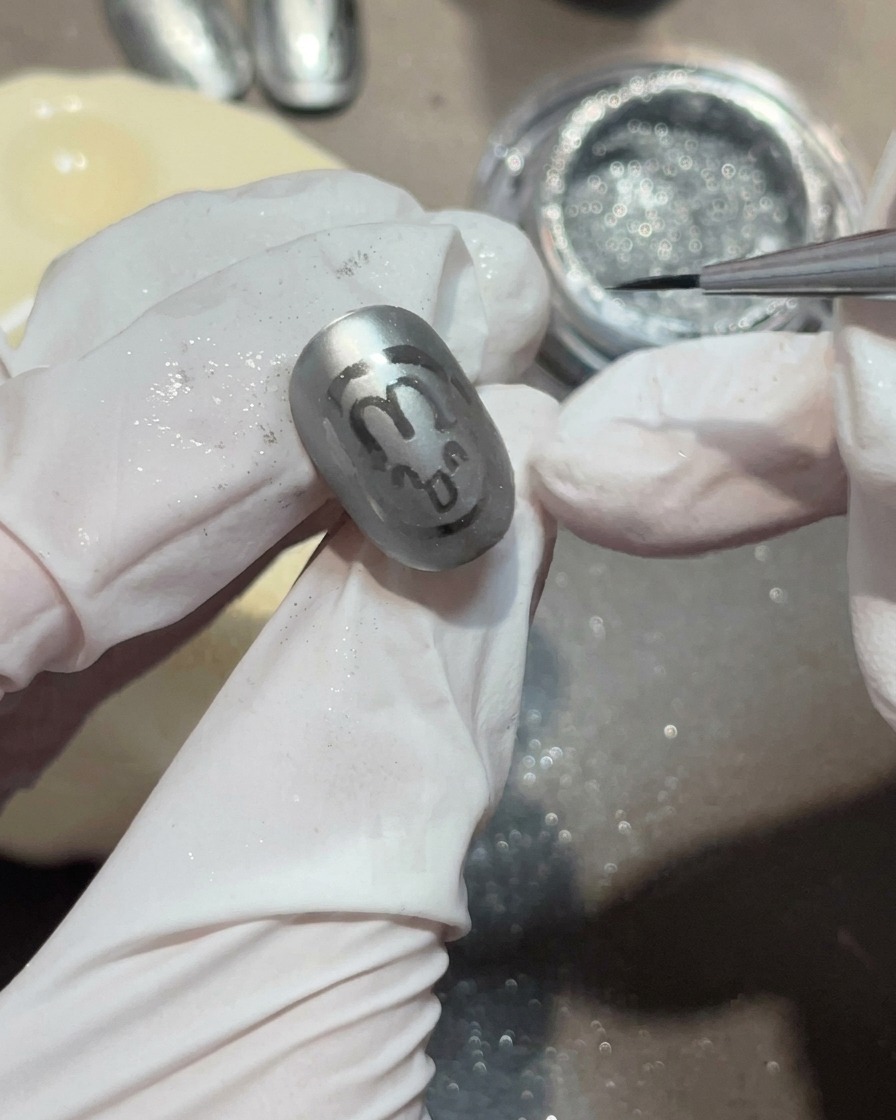 Primer plano de un pincel aplicando una pequeña gota de gel transparente sobre una uña gris mate para formar una gota de agua.