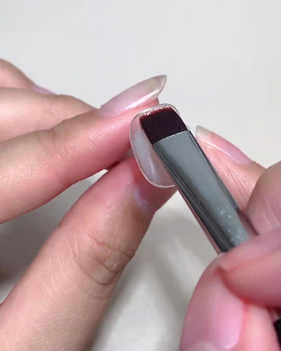 Un pincel aplicando una capa de gel cat eye nude sobre una uña con base curada.