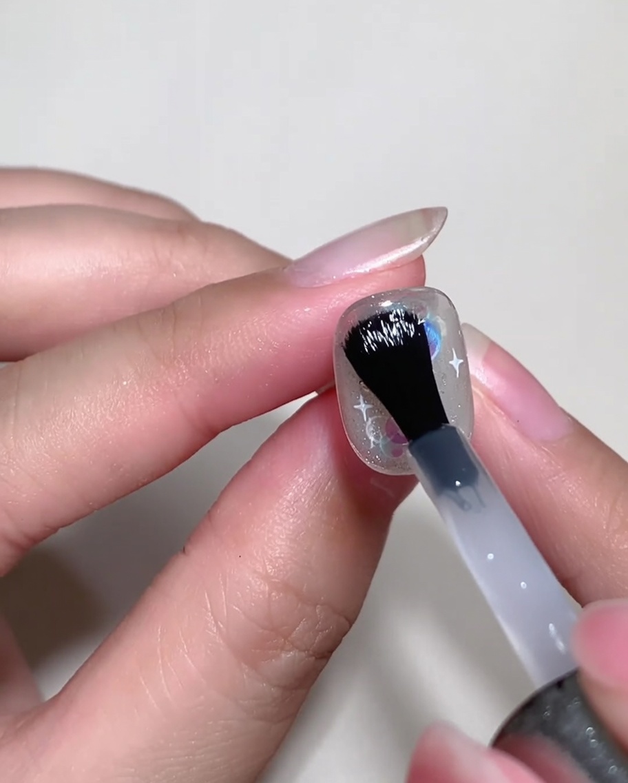 Un pincel de uñas aplicando un builder gel transparente sobre una uña decorada para nivelar la superficie.