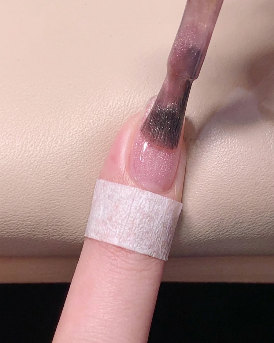 Vista superior mostrando un pincel nivelando el esmalte de gel rosa sobre la uña para un acabado uniforme.