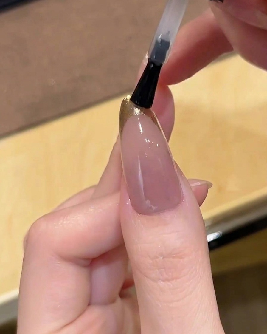 Un pincel aplicando un top coat transparente y brillante sobre una uña con punta francesa cromada dorada.