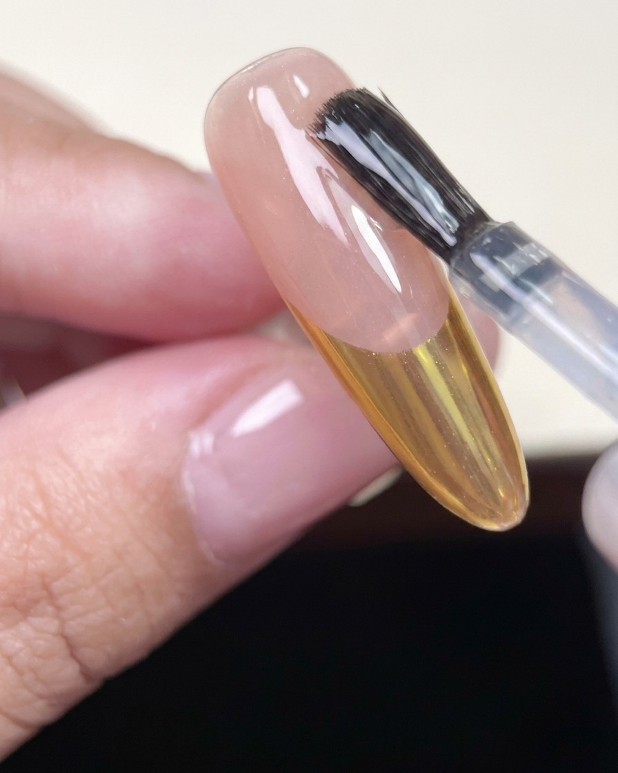 Pincel aplicando un top coat brillante sobre una uña con punta dorada.