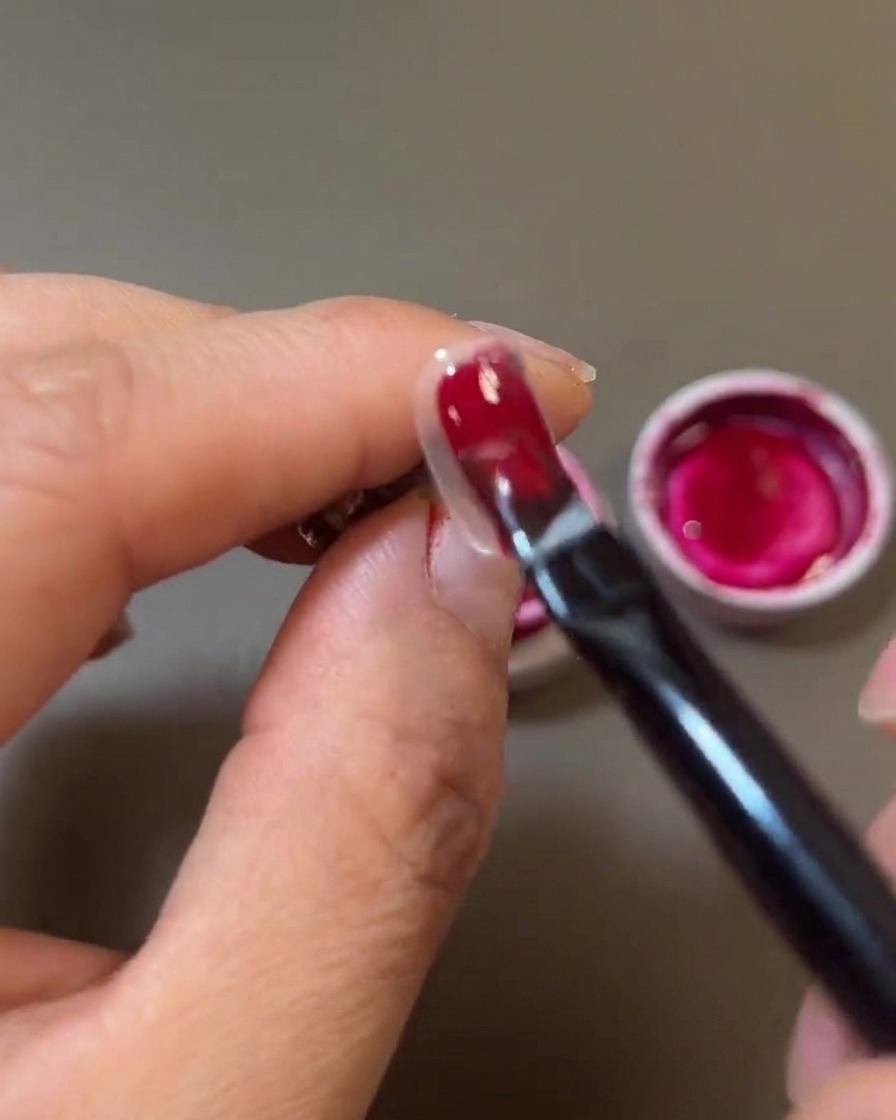 Primer plano de un pincel de uñas aplicando un gel rojo intenso sobre la superficie de la uña.