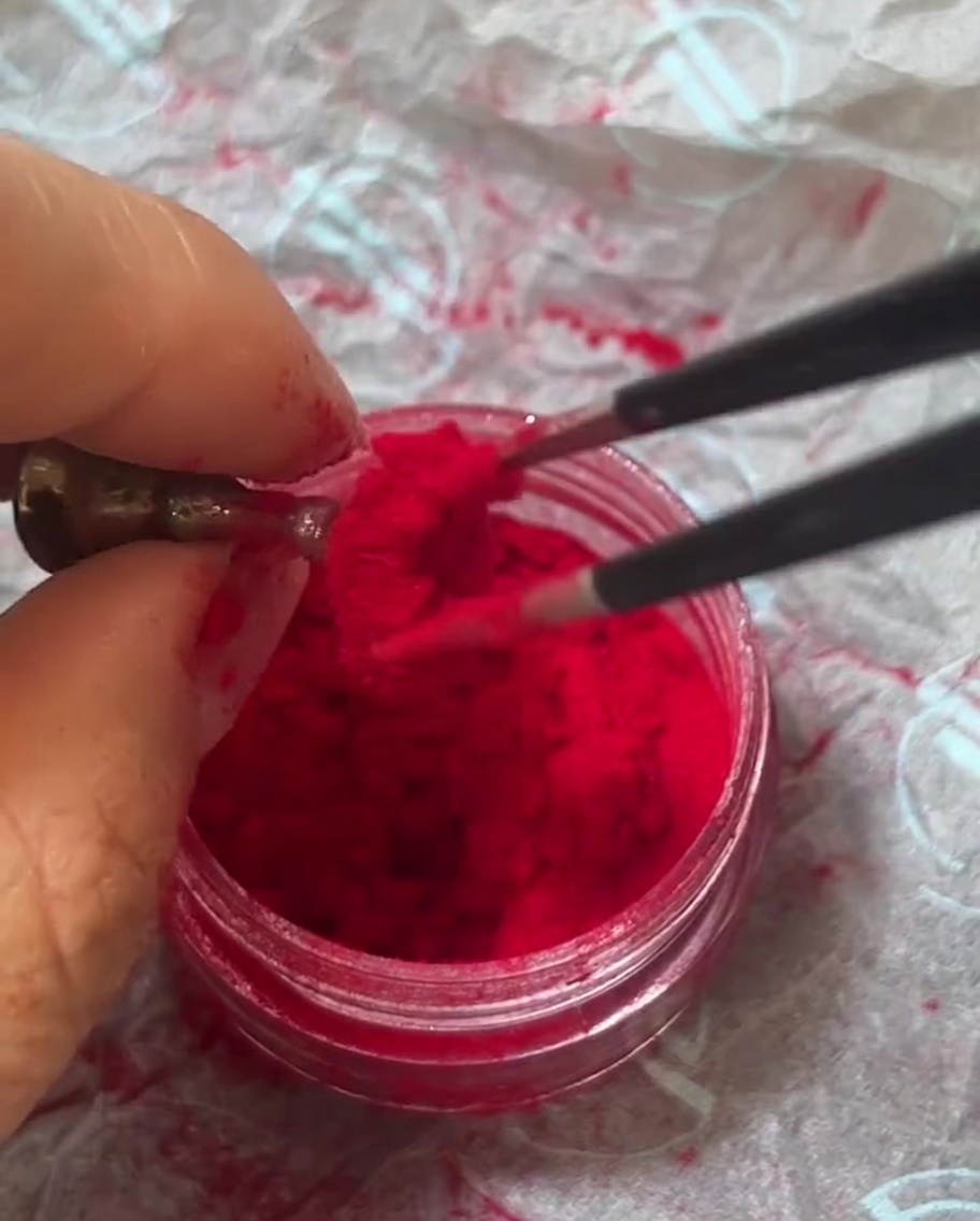 Pinzas metálicas sosteniendo un frasco de polvo de felpa rojo aterciopelado, recogiendo una pequeña cantidad de fibras.