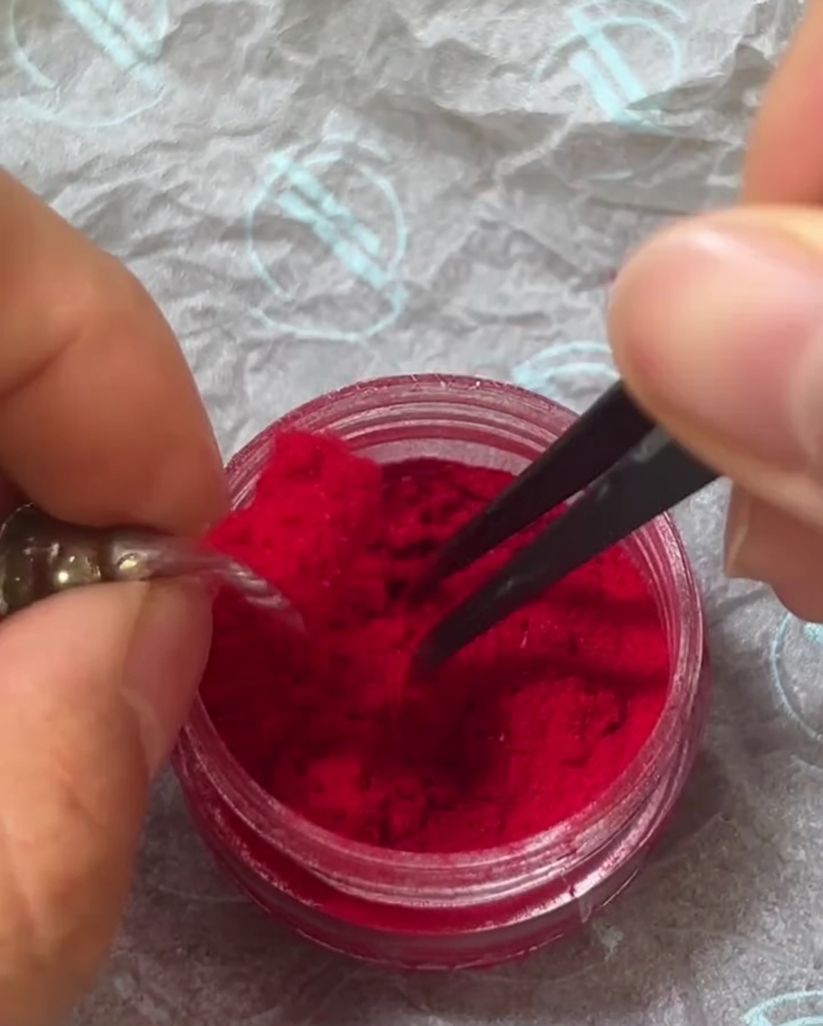 Pinzas espolvoreando cuidadosamente el polvo de felpa rojo sobre la superficie de una uña cubierta con gel húmedo.