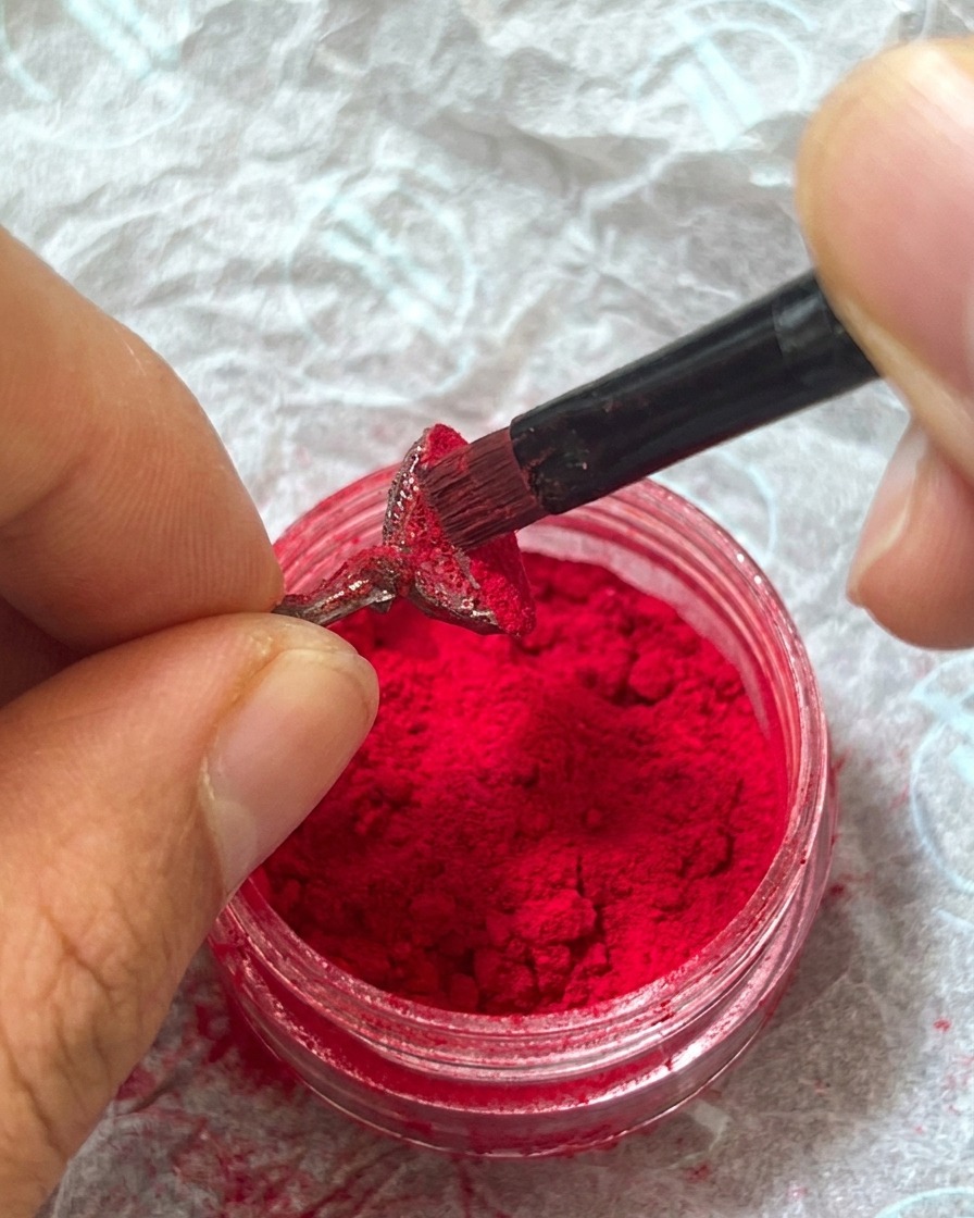 Un dedo presionando suavemente sobre el polvo de felpa rojo aplicado en la uña para fijarlo en el gel húmedo.