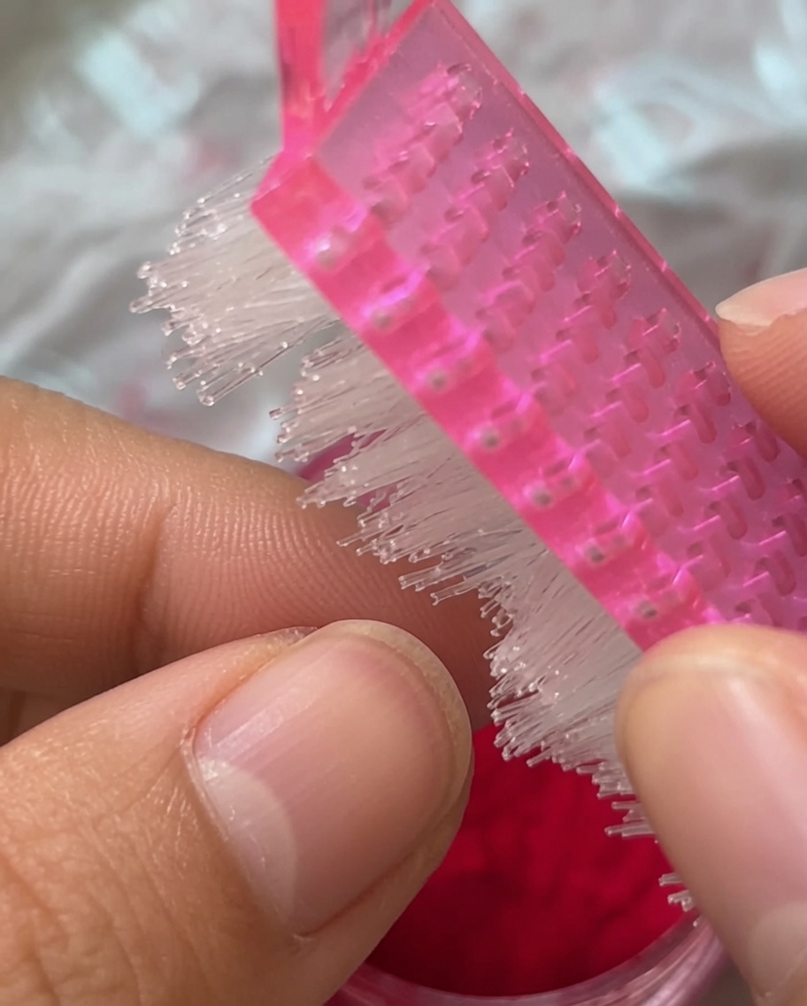 Primer plano de un cepillo para manicura rosa barriendo una uña con acabado de felpa roja.