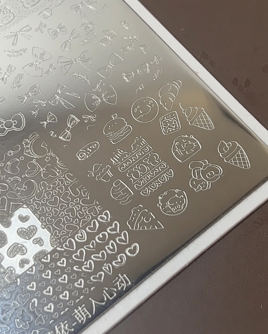 Un pincel de arte de uñas aplicando esmalte blanco espeso sobre íconos de comida grabados en una placa de estampado de metal.