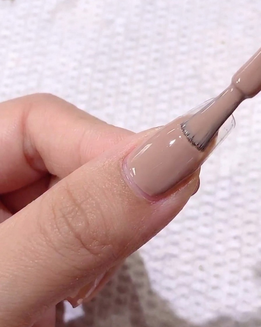 Primer plano de un pincel aplicando gel de color marrón claro sobre una uña natural preparada.