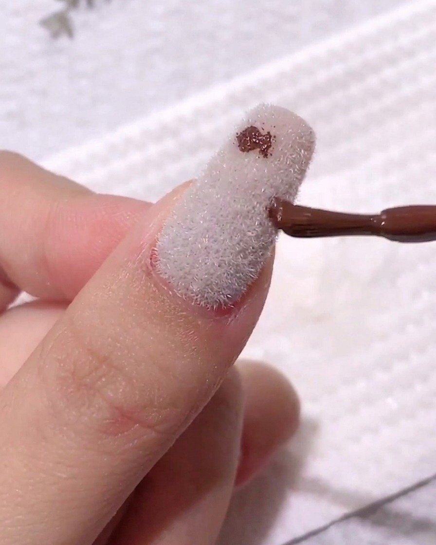 Pincel depositando puntos pequeños de gel color café oscuro sobre una superficie aterciopelada blanca.