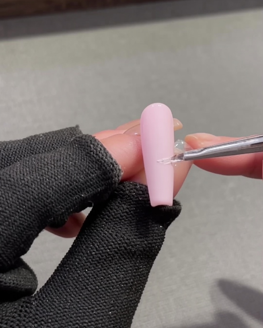 Un pincel de detalle fino aplicando gel relieve blanco texturizado para crear una línea de sonrisa francesa en una base de uña rosa.