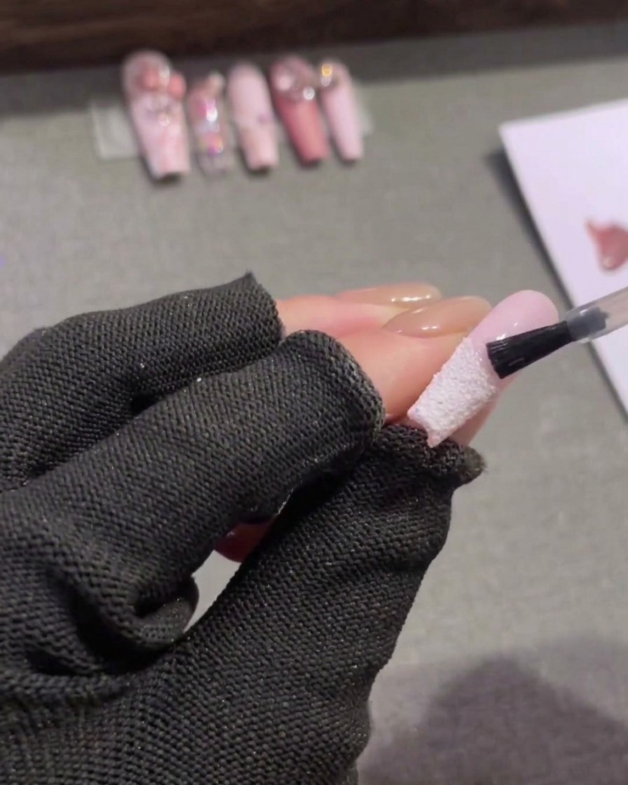 Un pequeño pincel de uñas aplicando un top coat mate transparente a la mitad superior rosa de una uña en forma de almendra, deteniéndose justo por encima de una punta francesa blanca texturizada.