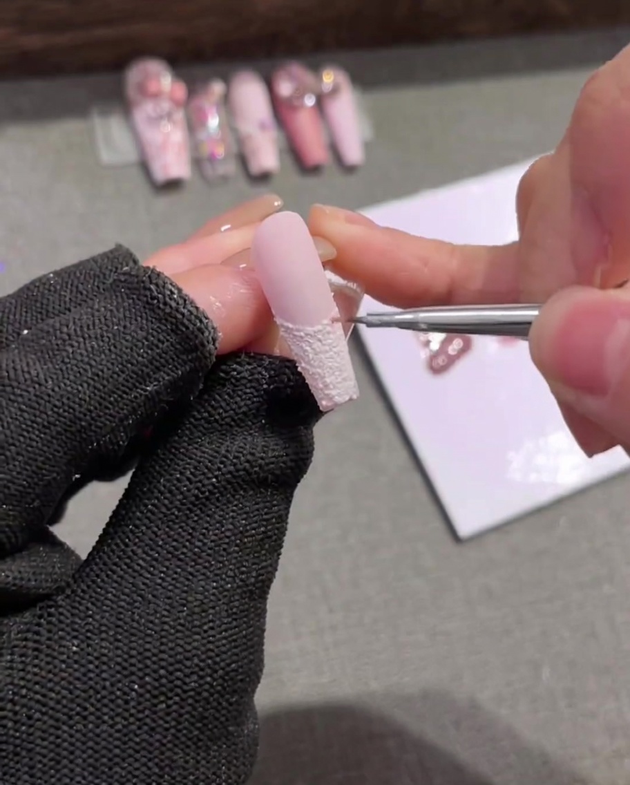 Un primer plano de un pincel liner aplicando delicadas rayas verticales de color rosa claro sobre una uña con punta francesa blanca texturizada.