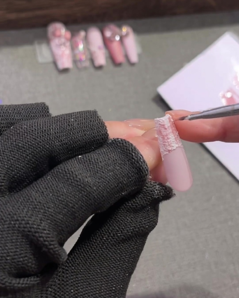 Un pincel de detalle para uñas añadiendo finas líneas marrones oscuras que se cruzan en un patrón de cuadros rosa y blanco.