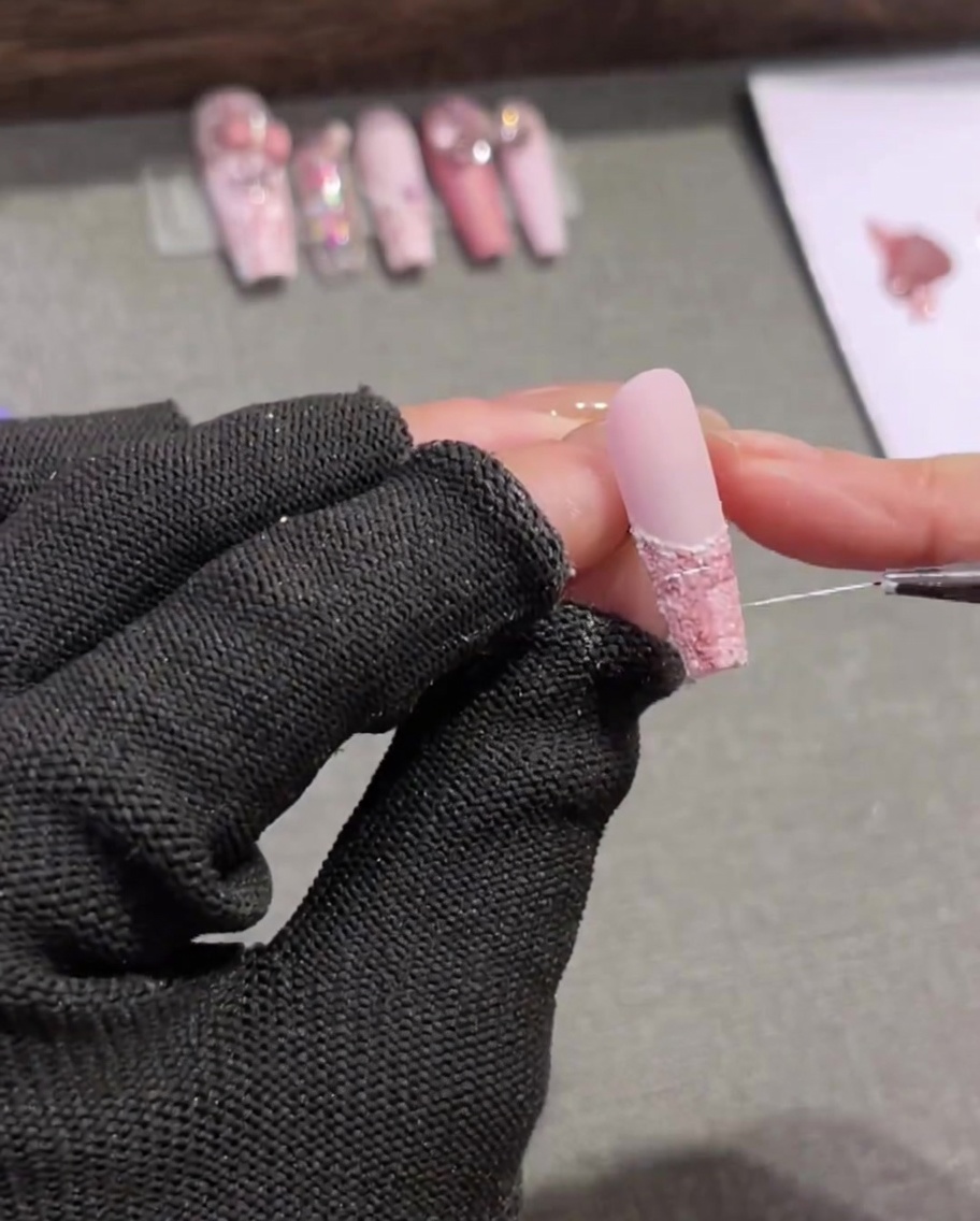 Un pincel liner muy fino y largo trazando delicadas líneas blancas aleatorias a través de un diseño de uñas tweed rosa y marrón.