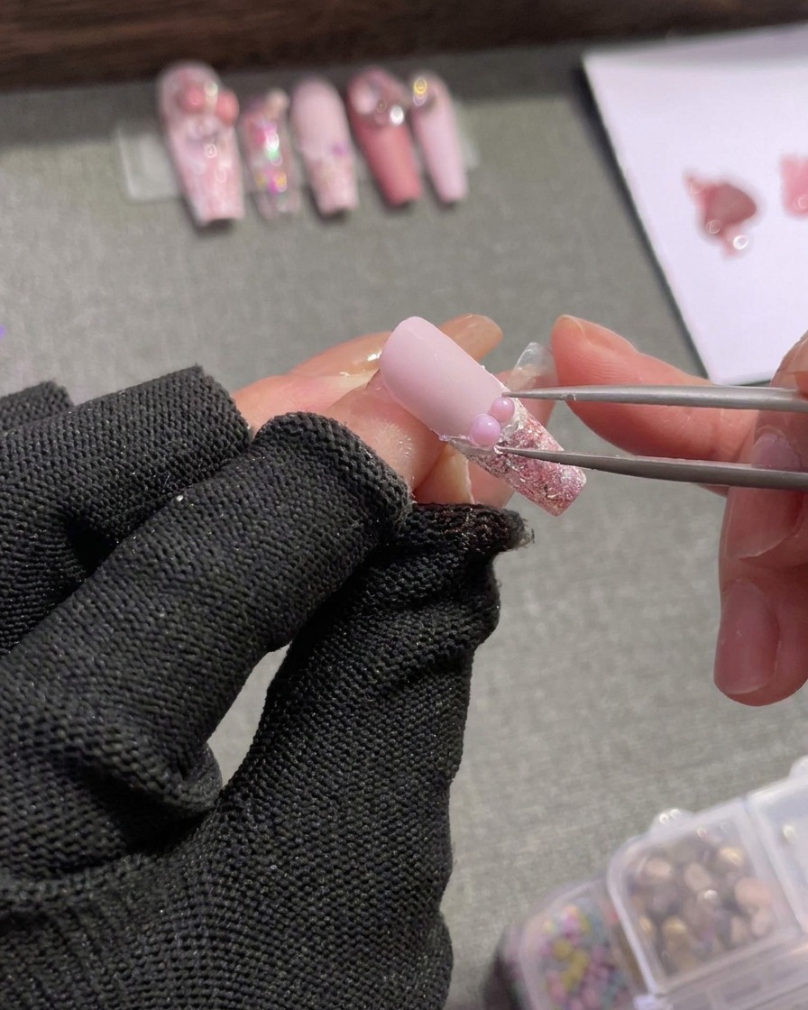 Pinzas posicionando cuidadosamente pequeños diamantes de imitación plateados y un pequeño charm de lazo rosa junto a una perla en el borde de la punta francesa.