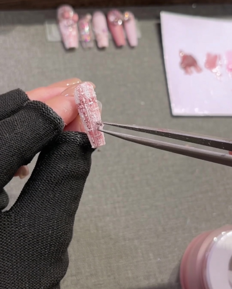 Un par de pinzas colocando pequeños diamantes de imitación decorativos sobre un tip de uña pintado a cuadros y texturizado.
