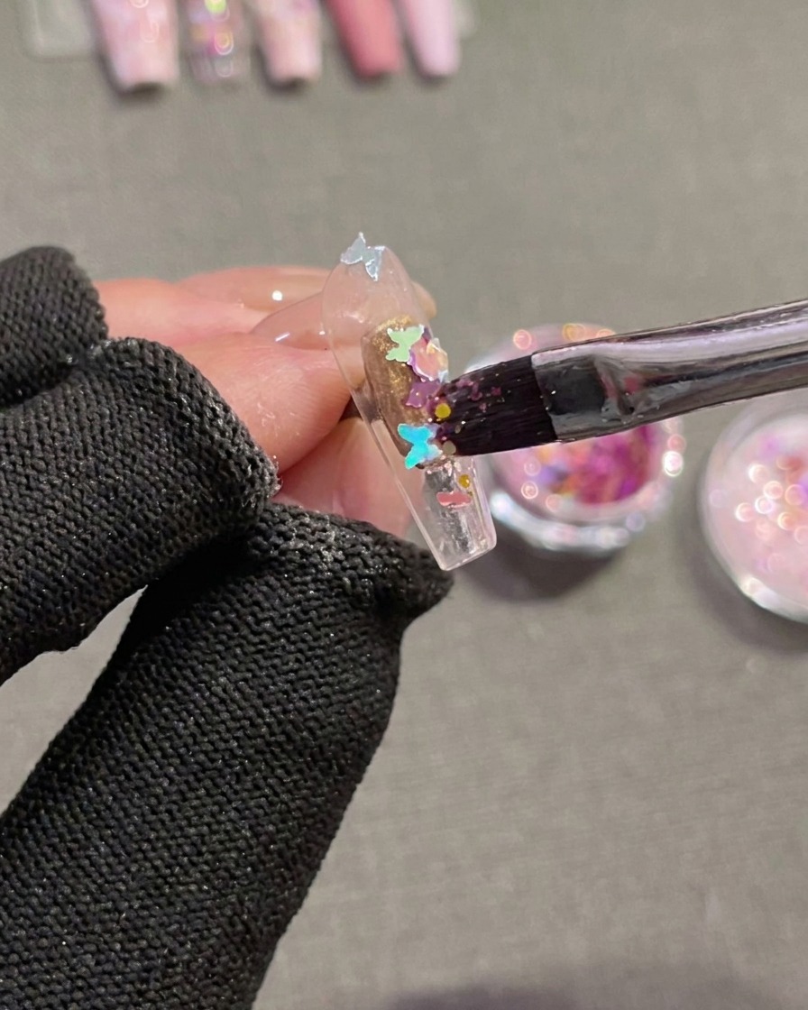 Una vista de primer plano de un pincel siendo usado para presionar lentejuelas holográficas en forma de mariposa sobre un tip de uña transparente recubierto con gel húmedo y sin curar.