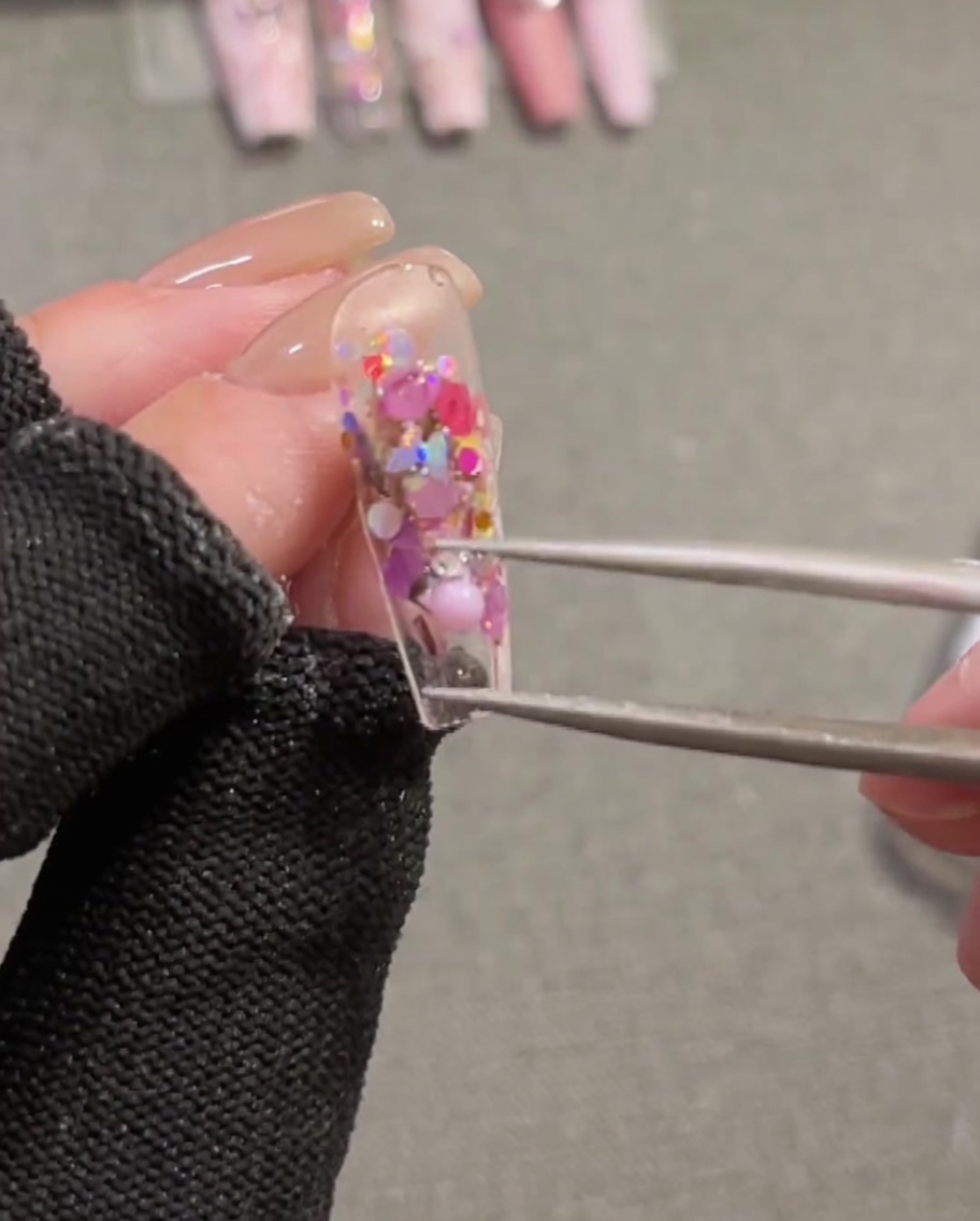 Primer plano de pinzas sosteniendo y posicionando un charm de lazo 3D de cinta rosa sobre una uña que tiene gel constructor húmedo.