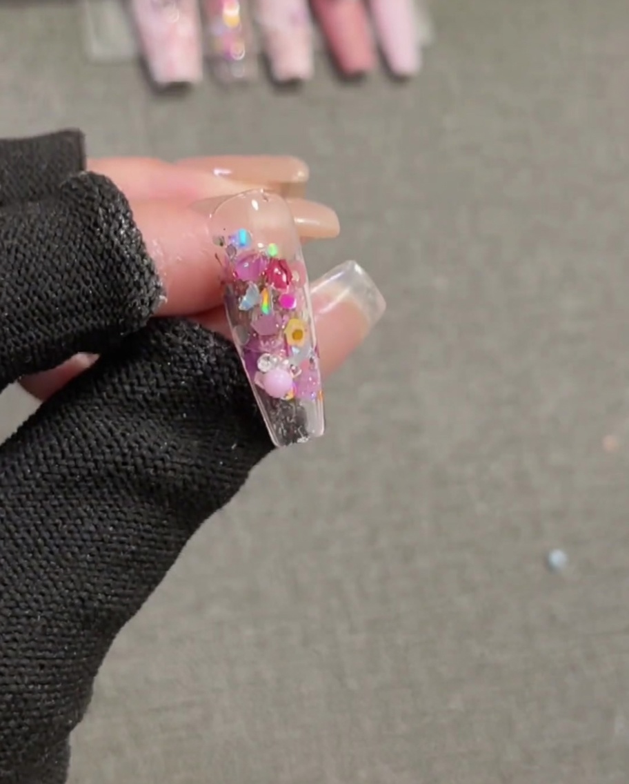Un pincel aplicando una capa final de top coat transparente de alto brillo sobre una uña completa que presenta lentejuelas incrustadas y un charm de lazo rosa 3D.