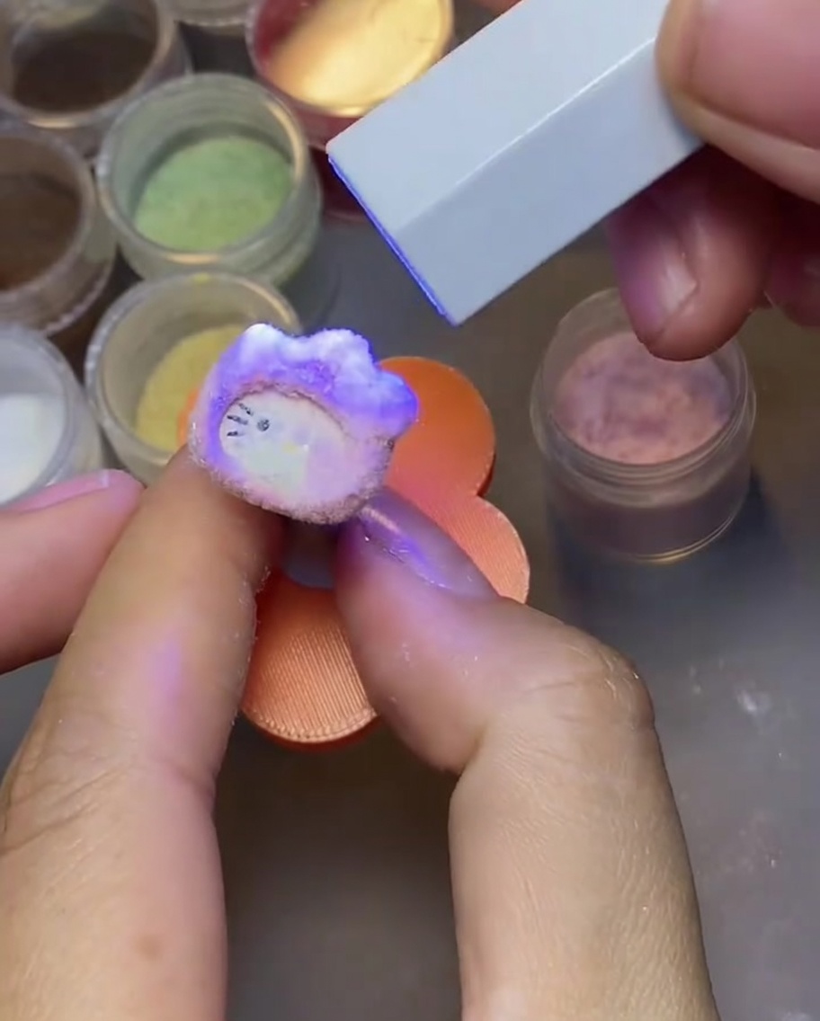 Una mano dando golpecitos a un dije de uña de Hello Kitty curado para eliminar el exceso de polvo de terciopelo.