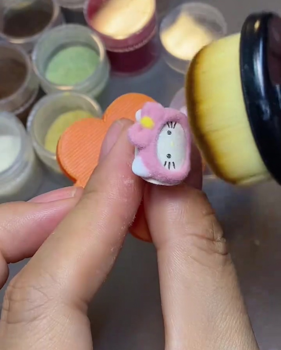 Un primer plano de un pincel de maquillaje esponjoso barriendo el exceso de polvo de terciopelo rosa de la superficie de un dije de uña de Hello Kitty.