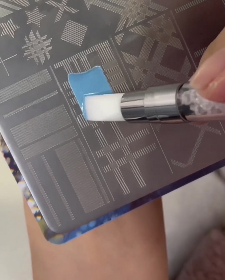 Un pincel aplicando esmalte azul claro sobre una sección con patrón de cuadros en una placa de metal para estampar uñas.
