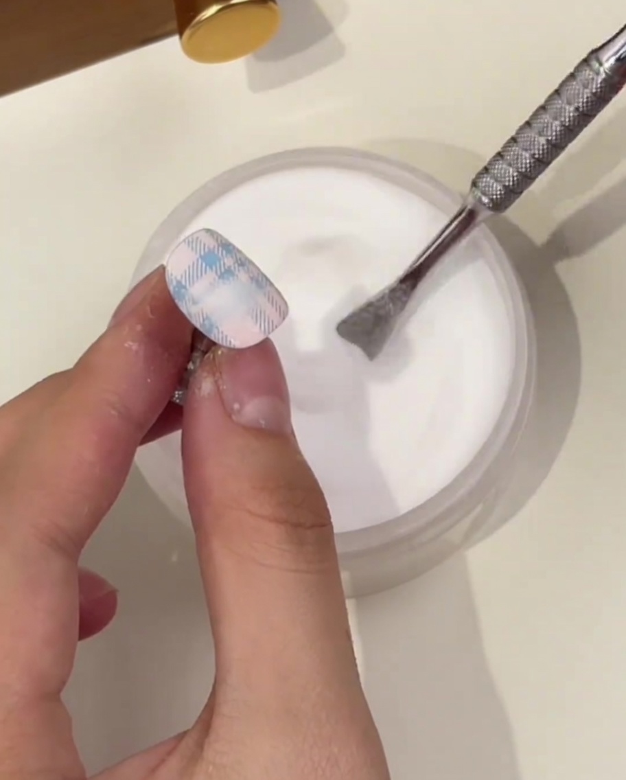 Un primer plano de una mano sosteniendo una uña con un patrón de cuadros azules, usando un empujador de cutículas para espolvorear polvo transparente sobre el diseño húmedo.
