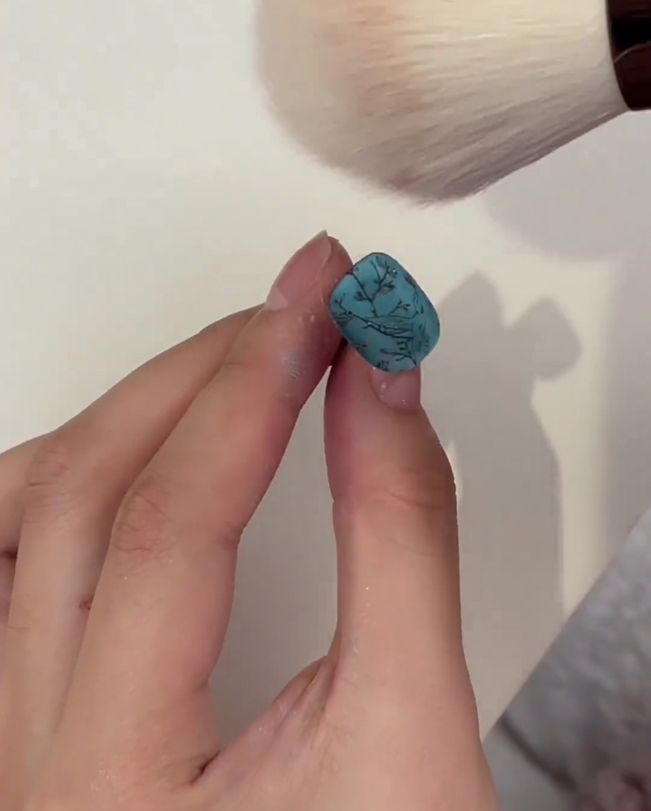 Una brocha estilo maquillaje esponjosa quitando el polvo restante de una uña turquesa después del curado.