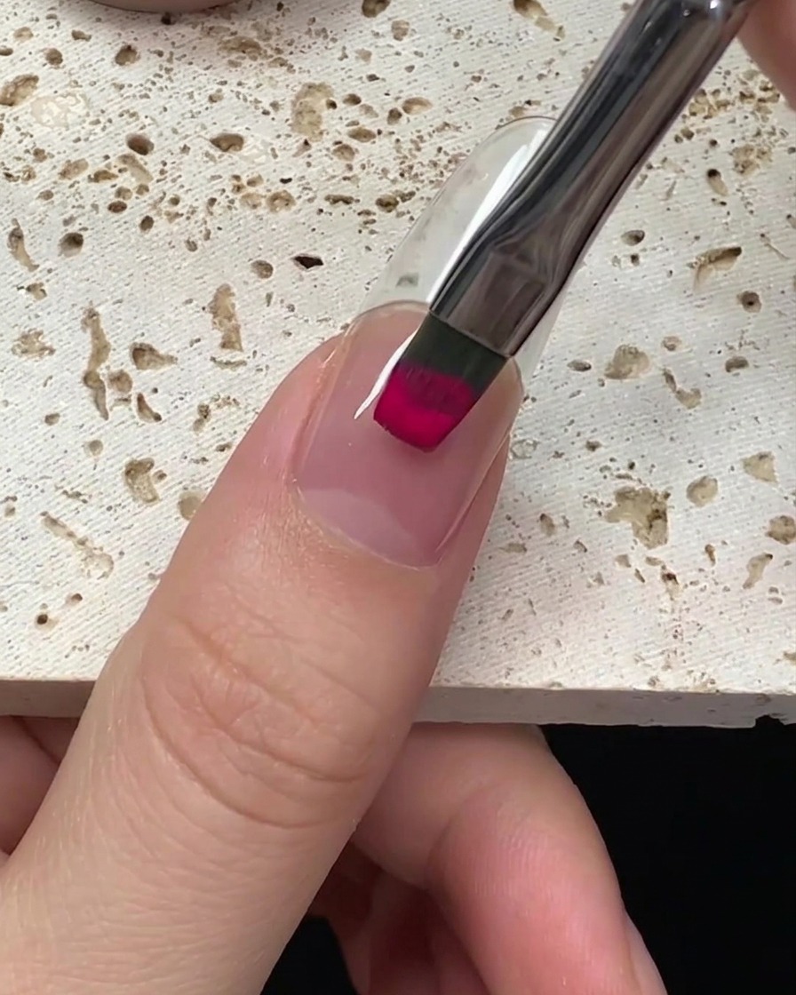 Vista cercana de un pincel aplicando esmalte de gel magenta vibrante en una uña, comenzando desde la cutícula.