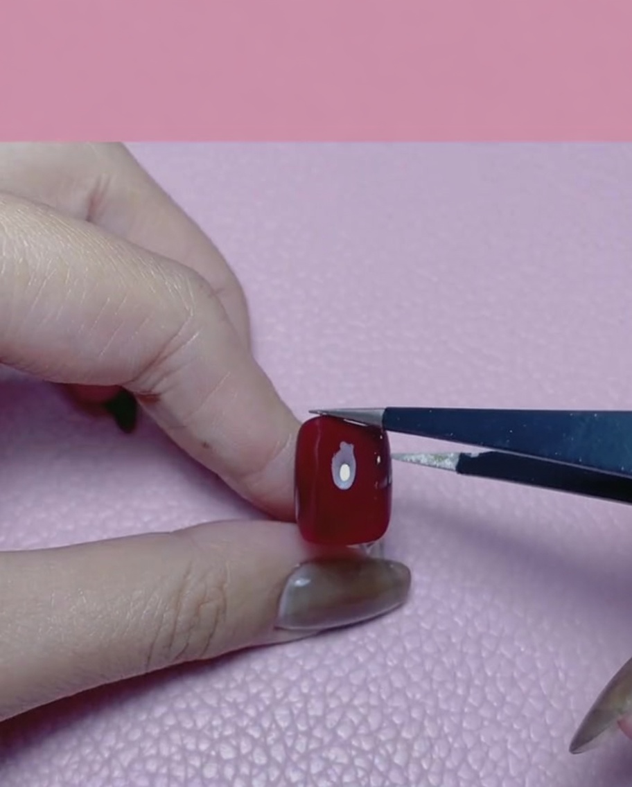Una mano usando pinzas para aplicar un trozo de pan de oro sobre una uña pintada de rojo brillante.