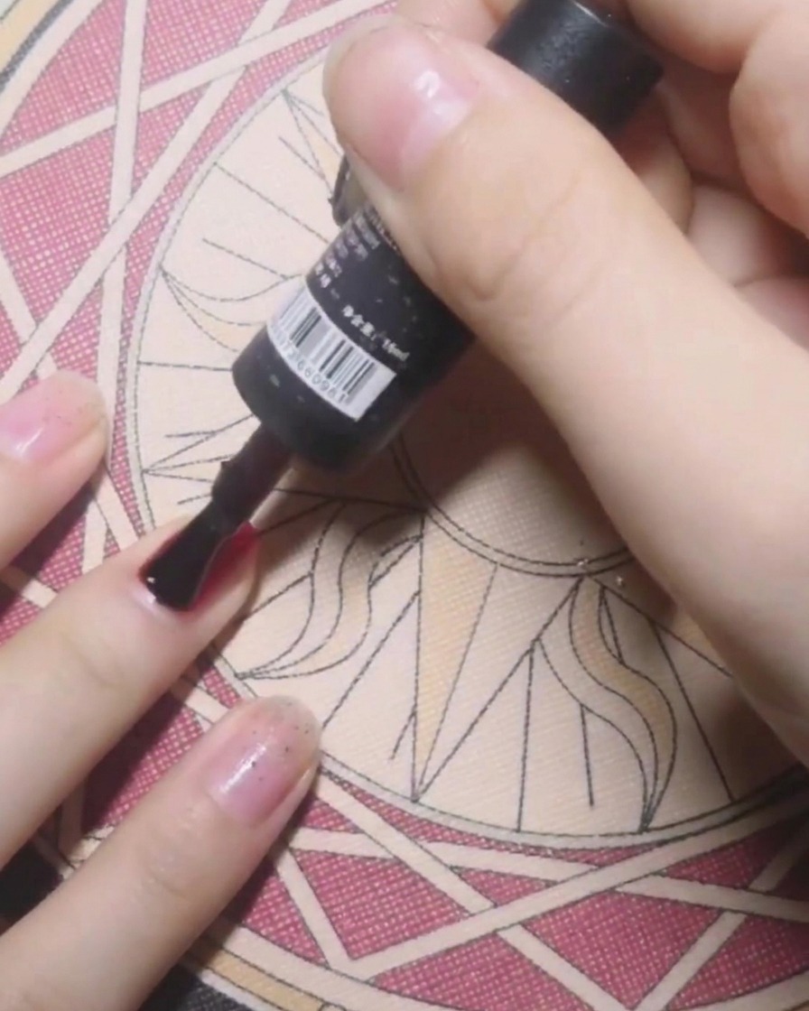 Un pincel de botella aplicando base coat transparente sobre una uña en preparación para esmalte de gel rojo sólido.