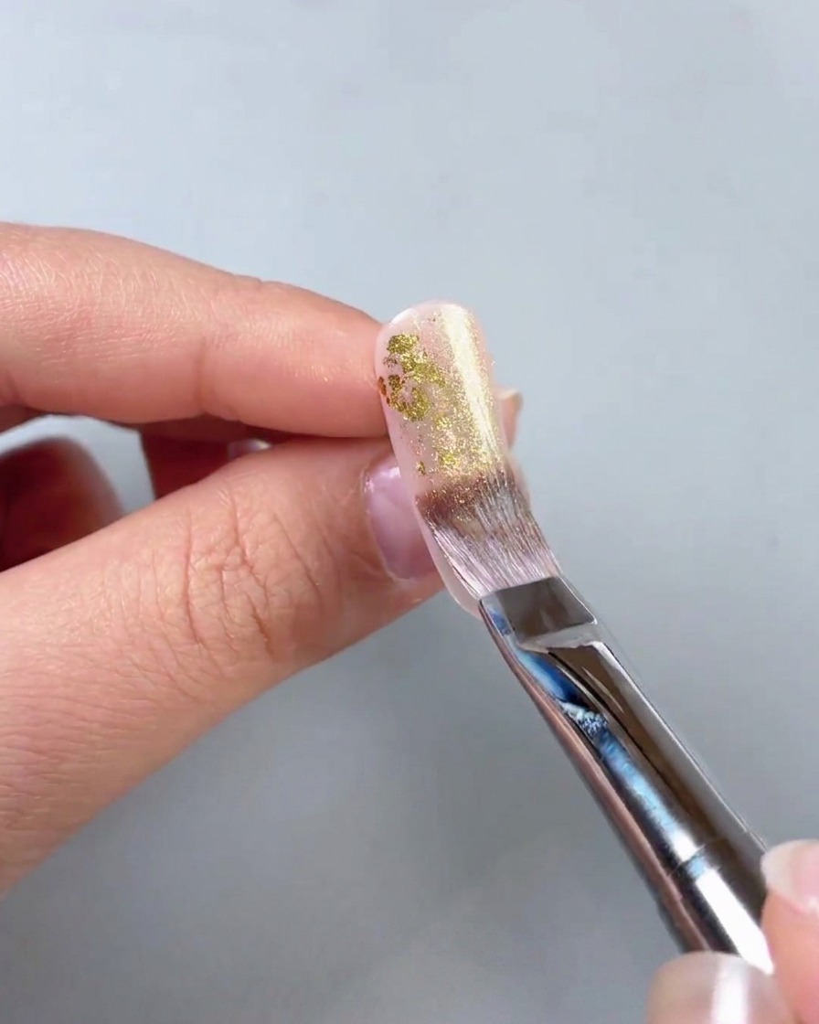 Primer plano de un pincel aplicando gel dorado brillante sobre piezas de lámina de oro en una uña.