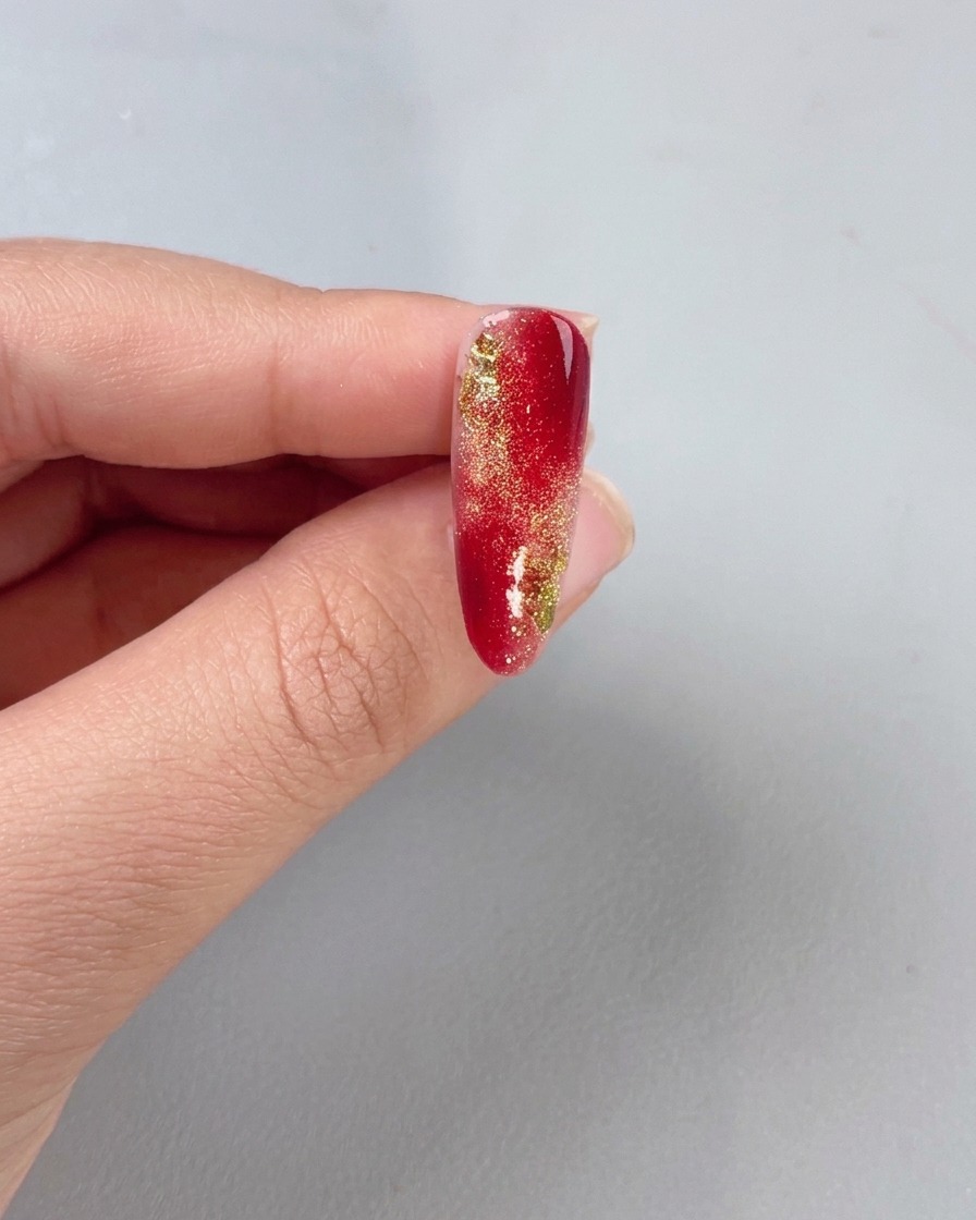 Un pincel aplicando un top coat transparente sobre una uña con degradado rojo y oro.