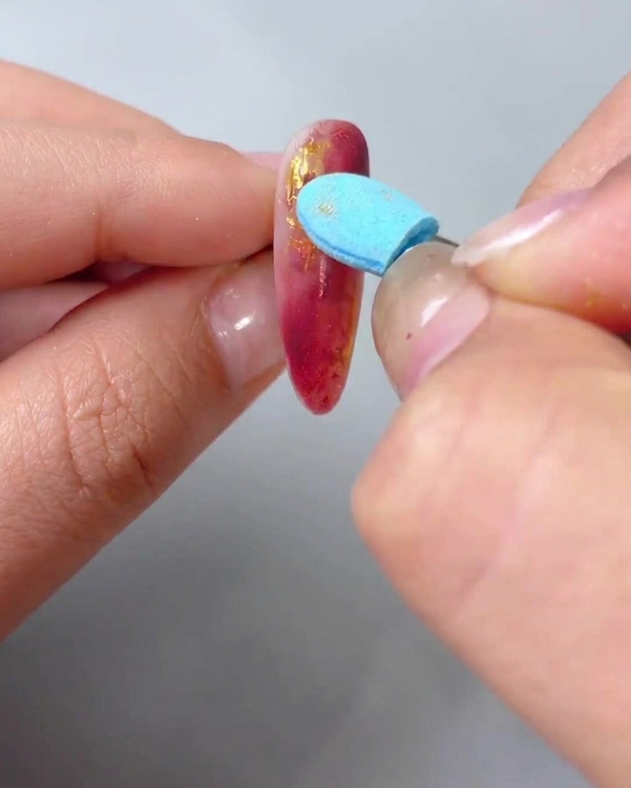 Primer plano de una mano usando una esponja aplicadora azul para frotar polvo de oro metálico sobre las líneas de textura de una uña de mármol rojo.