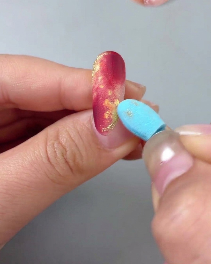 Primer plano de una mano con una uña de mármol rojo y oro, mientras una herramienta de esponja azul retira suavemente el exceso de polvo de oro.
