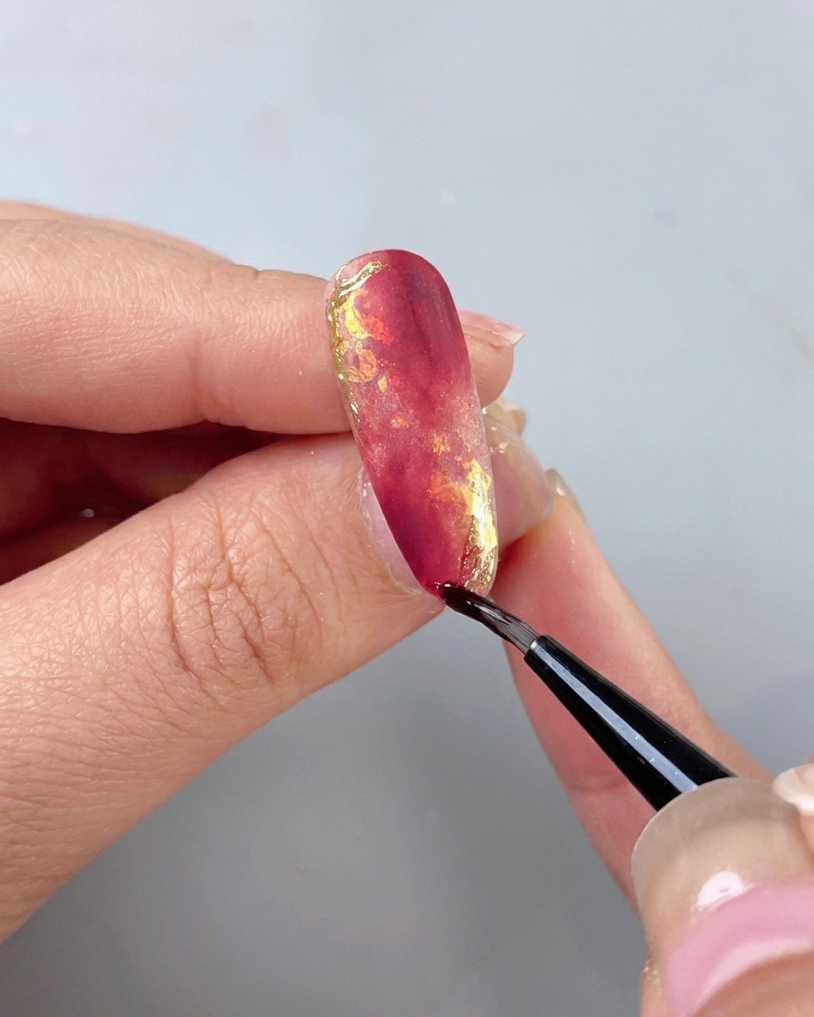 Un pincel fino de detalle aplicando un top coat aislante transparente sobre una uña de mármol rojo y oro.