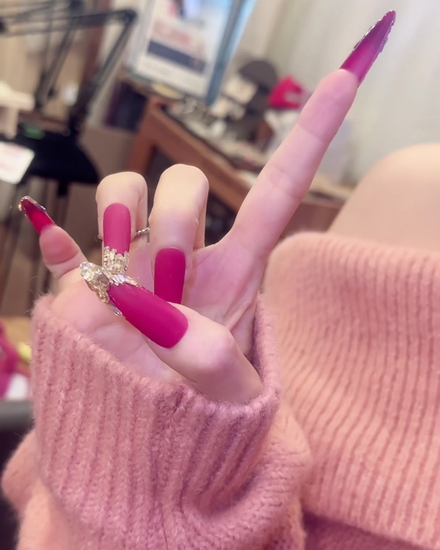 Una mano con los dedos extendidos, mostrando uñas largas rosas mate con acentos de cristal junto a elegantes accesorios de anillos plateados.