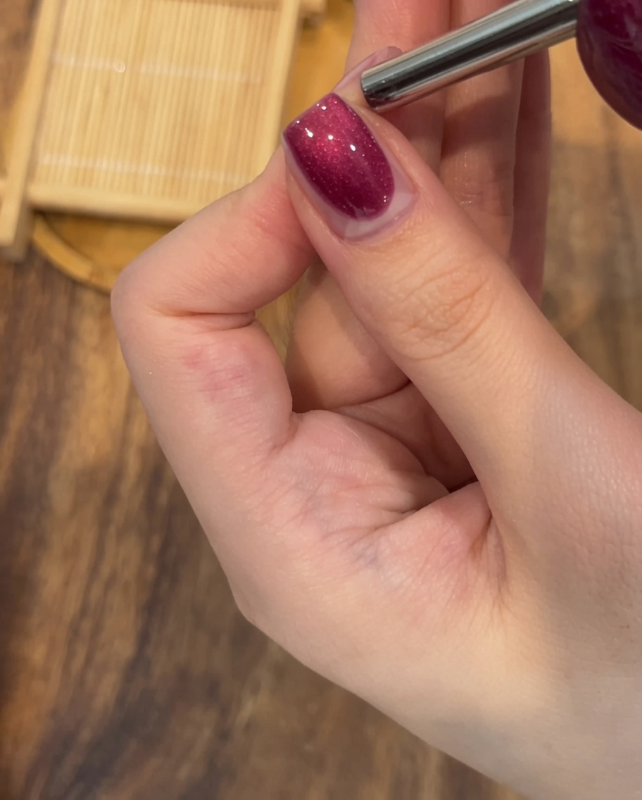 Primer plano de un imán rectangular sostenido firmemente sobre una uña pintada con gel magnético húmedo para crear una línea de brillo prominente.