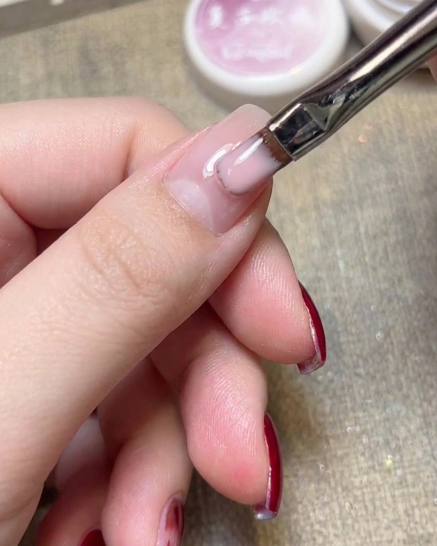 Primer plano de un pincel plano para gel aplicando esmalte jelly rosa claro sobre una uña natural.