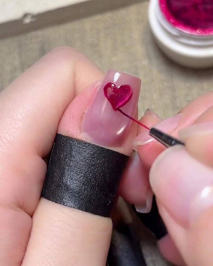 Un pincel delineador fino pintando cuidadosamente un corazón magenta sobre una uña rosa claro.