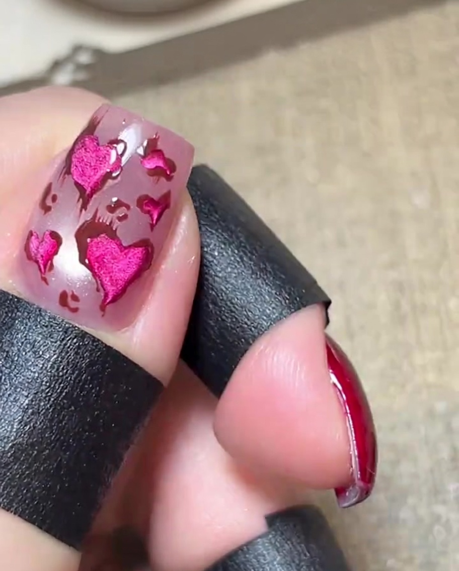 Primer plano de una uña rosa brillante con corazones rosas metálicos delineados en marrón, con líneas marrones finas y delicadas que se extienden hacia afuera de los diseños.