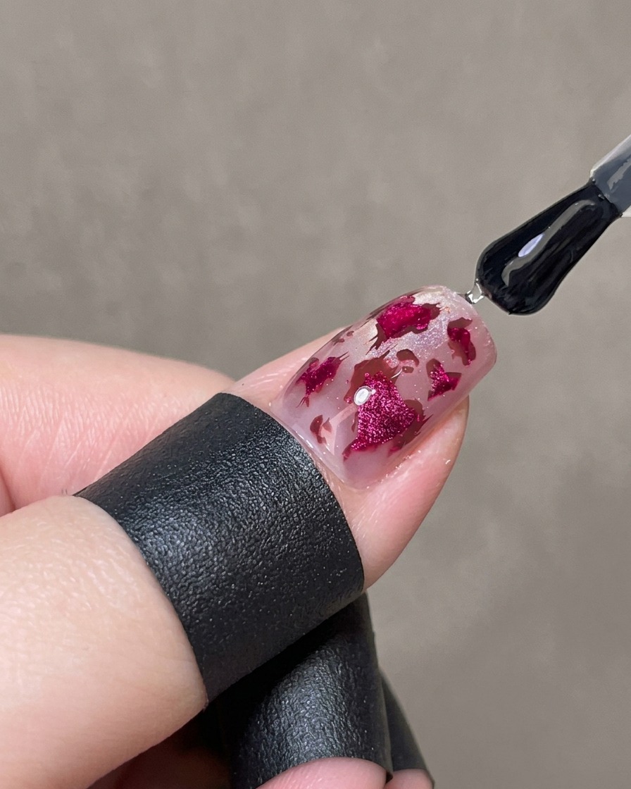 Un pincel aplicando una capa gruesa de gel transparente brillante sobre una uña decorada para encapsular el diseño de corazones rosas y manchas de leopardo.