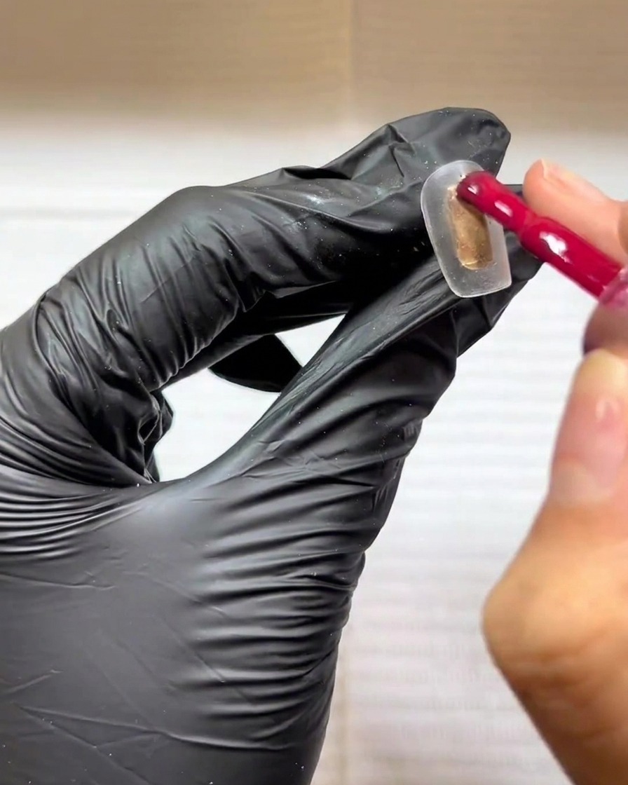 Mano enguantada aplicando gel rosa rojizo sobre un tip transparente.